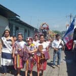 Niñas y jóvenes en una celebración tradicional de Guatemala con trajes típicos y bandera en mano, participando en un desfile cultural y festivo en la calle. – SoyMigrante.com REVISTA