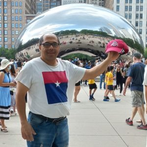 Atractivo hombre en un evento en Chicago tocando la popular escultura Cloud Gate, también conocida como "The Bean", con multitudes y edificios altos en el fondo, promoviendo cultura y comunidad.