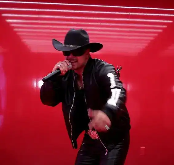 Estilo musical mexicano, cantante con sombrero negro y gafas de sol en escenario rojo, presentación de música regional, artista popular en México.
