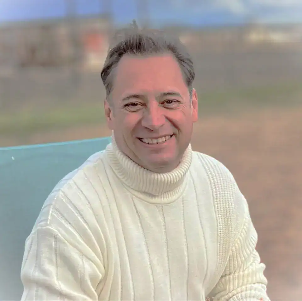 Hombre sonriendo con suéter gris claro en un entorno al aire libre. – SoyMigrante.com REVISTA