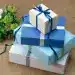 Paquete de regalo envuelto con papel azul y blanco, decorado con lazo azul, sobre una mesa de yute, ideales para ocasiones especiales y celebraciones con temática de migrantes y comunidad.