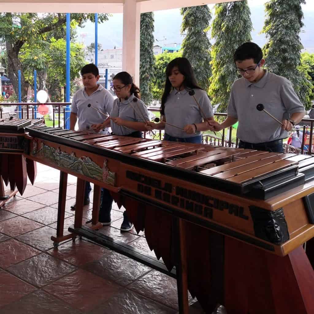 Niños tocando xilófono en evento escolar en un espacio abierto con flores y árboles. – SoyMigrante.com REVISTA