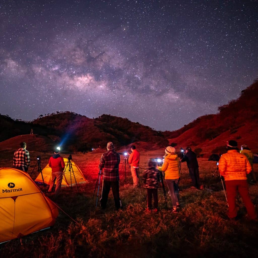 Observatorio astronómico nocturno en un paisaje montañoso con campamentos y telescopios, bajo un cielo lleno de estrellas y la Vía Láctea, en un evento de astronomía y ciencia para migrantes. – SoyMigrante.com REVISTA