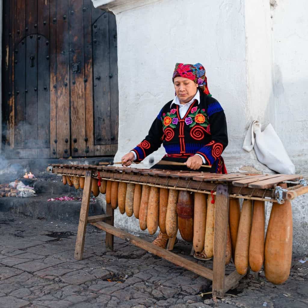 Mujer indígena tocando la flauta de pan tradicional en una calle de comunidad rural en México, mostrando cultura y identidad artesanal.