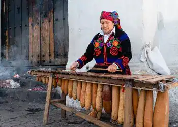 Mujer indígena tocando la flauta de pan tradicional en una calle de comunidad rural en México, mostrando cultura y identidad artesanal.