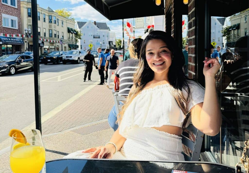 Ambiente urbano y multicultural en EE. UU., joven latina disfrutando de un día soleado en una terraza de café con una bebida refrescante, reflejando la experiencia de inmigrantes y la vida en el extranjero.