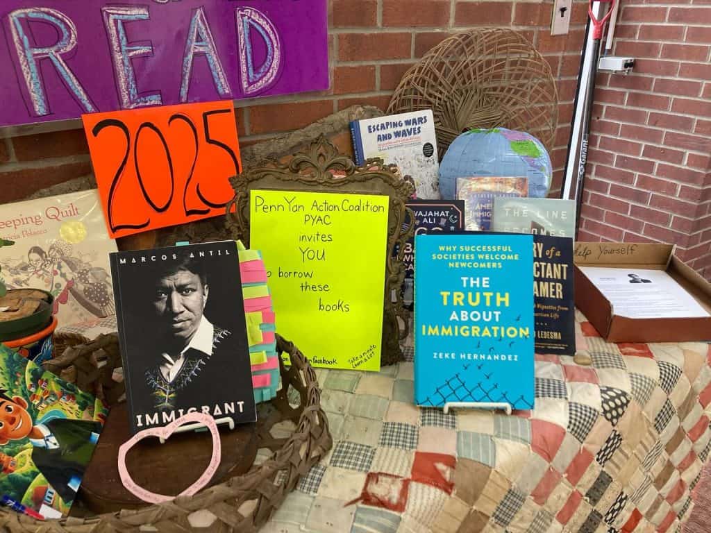 Migrante figura dentro de las lecturas del años 2025 en la Biblioteca y la Coalición de Penn Yan – SoyMigrante.com REVISTA Pasaporte y libros sobre inmigración y derechos de los inmigrantes en un puesto de lectura en la comunidad. – SoyMigrante.com REVISTA