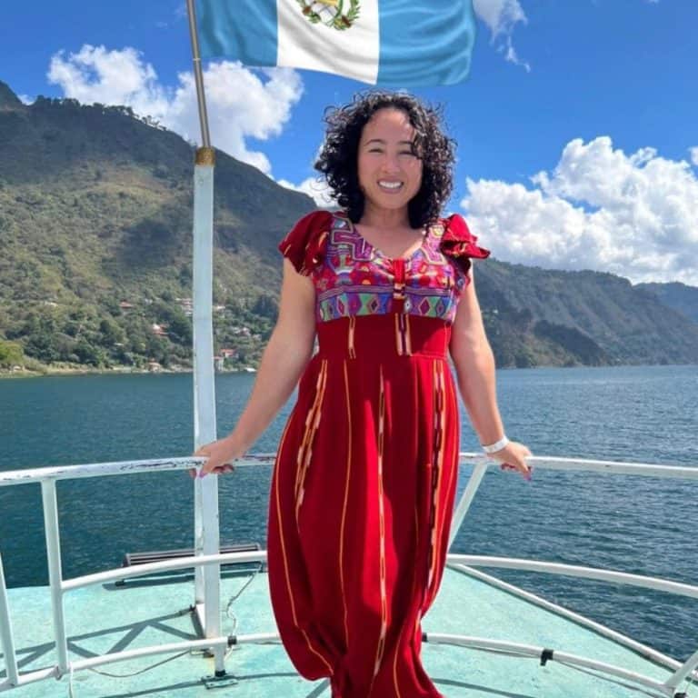 Mujer en una embarcación con bandera de Guatemala, paisaje montañoso y río, cultura migrante, orgullo nacional, turismo en Guatemala, paisaje natural, mujer sonriente, vestimenta tradicional, viaje y aventura, revista SoyMigrante.