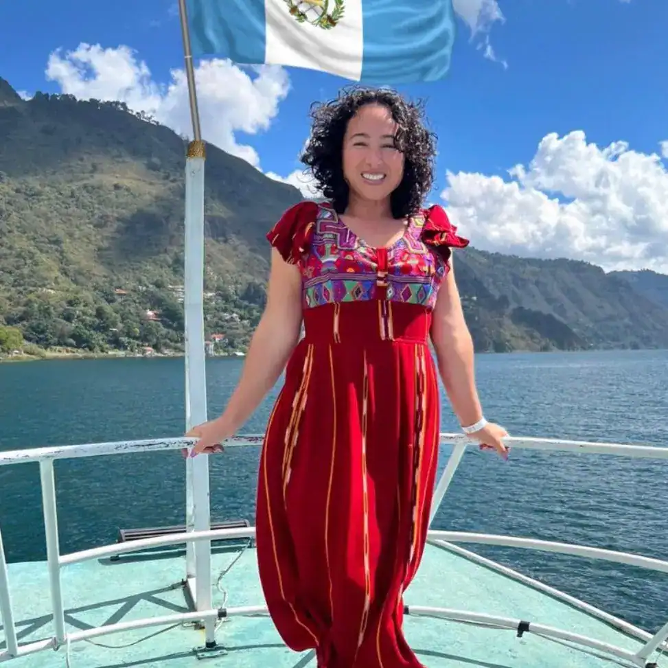 lissett garcia migrante guatemalteca en florida – SoyMigrante.com REVISTA Mujer en una embarcación con bandera de Guatemala, paisaje montañoso y río, cultura migrante, orgullo nacional, turismo en Guatemala, paisaje natural, mujer sonriente, vestimenta tradicional, viaje y aventura, revista SoyMigrante. – SoyMigrante.com REVISTA
