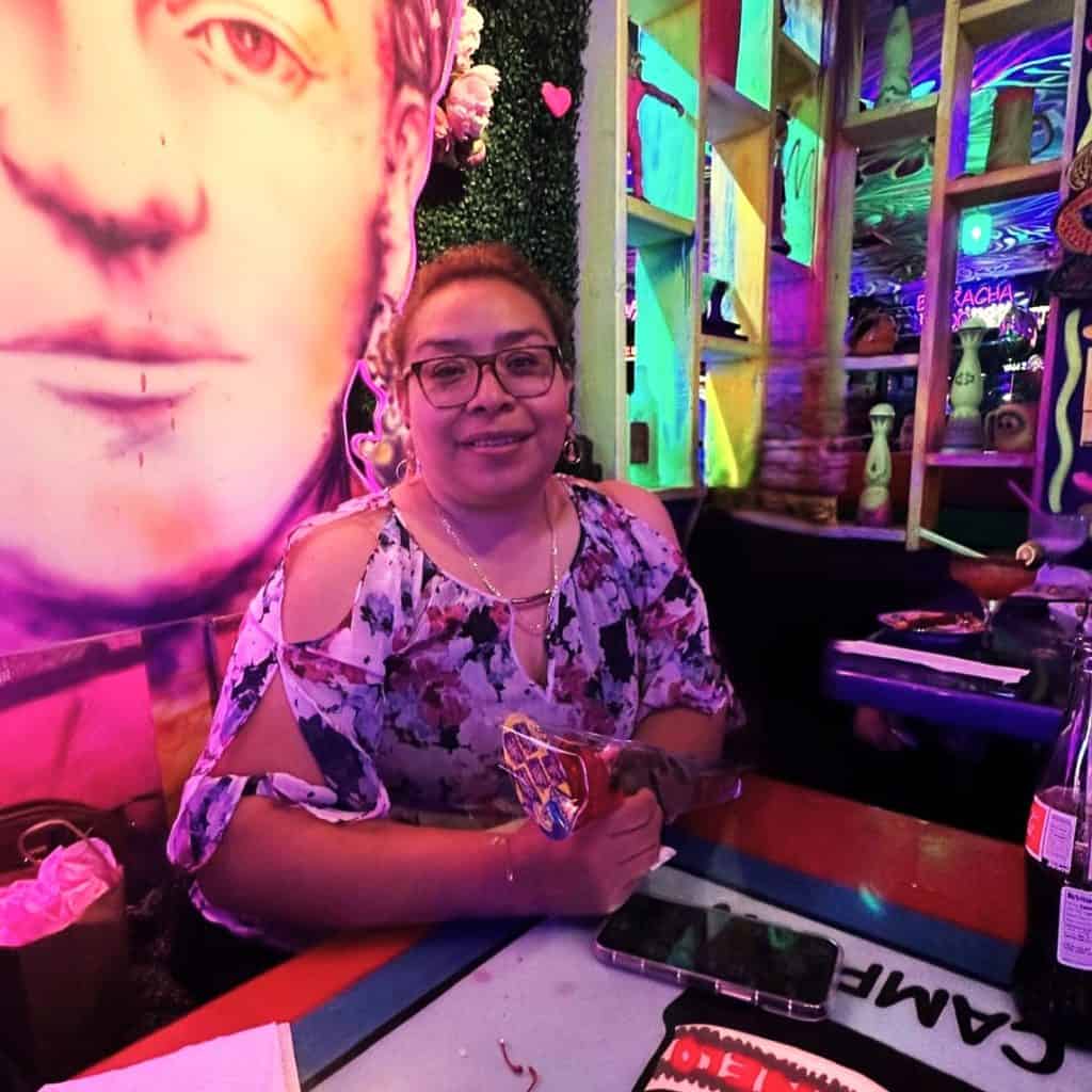 Disfrutando de la cultura mexicana en un ambiente vibrante con arte y color en SoyMigrante.com REVISTA. Mujer feliz en un restaurante con decoración artística y luces neón, celebrando la diversidad y tradiciones mexicanas. – SoyMigrante.com REVISTA