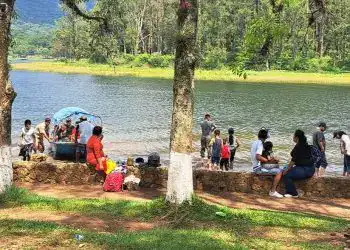 Personas disfrutando y socializando en un entorno natural junto a un lago en un día soleado, con árboles verdes y paisaje montañoso de fondo.