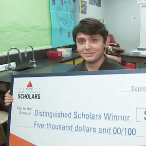 Premio de beca para estudiantes distinguidos, joven sosteniendo un gran cheque en un aula escolar. – SoyMigrante.com REVISTA