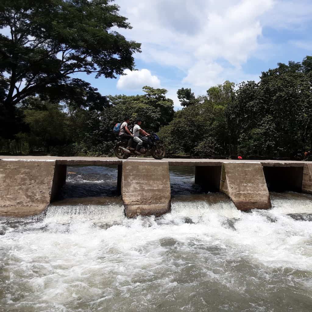 Moto en un puente sobre un río rodeado de árboles en un día soleado.