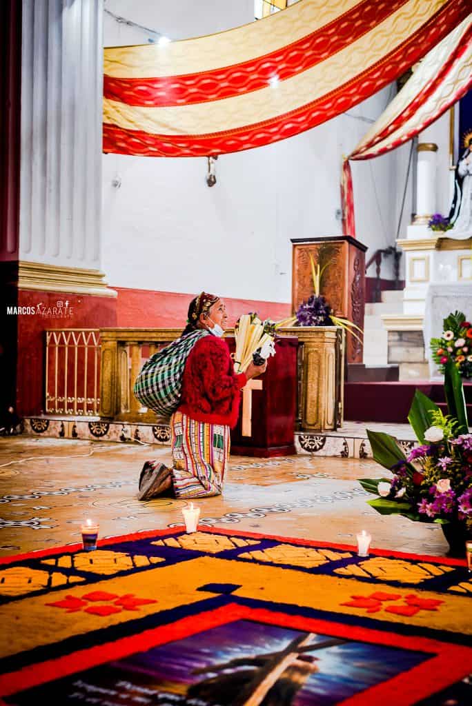 No hay oración pequeña ni plegaria que no sea escuchada en el Día de la Misericordia. – SoyMigrante.com REVISTA Mujer indígena en postura de oración en interior de iglesia durante ceremonia religiosa, con flores y velas. – SoyMigrante.com REVISTA