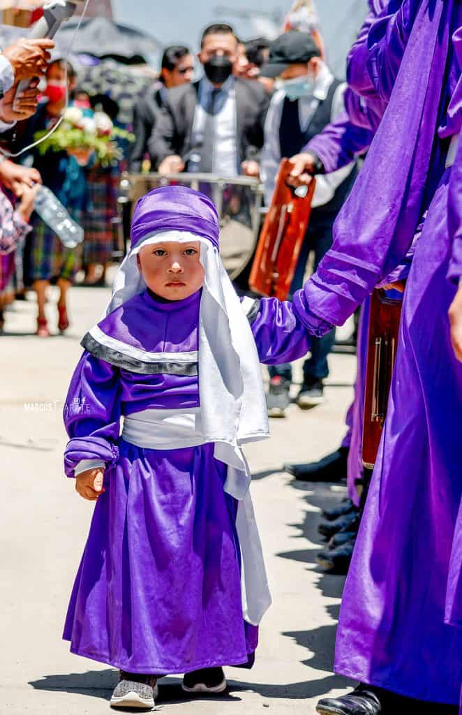 Y el hijo acompaña a su papá por el recorrido, porque es así como se siembra la semilla de la tradición. – SoyMigrante.com REVISTA Niña con vestimenta tradicional en celebración cultural, participando en una festividad con múltiples personas en el fondo. – SoyMigrante.com REVISTA