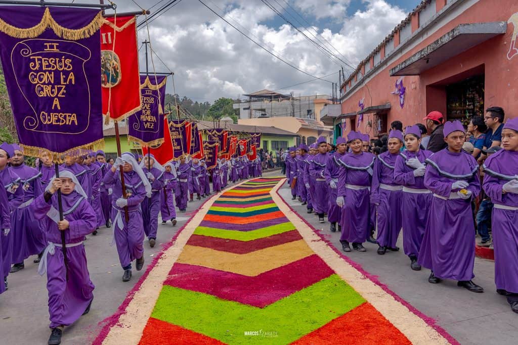 Los cucuruchos abarrotan las calles que parecen decir ¡Camina! sin usar una sola palabra. – SoyMigrante.com REVISTA Marcha de celebración religiosa con niños y jóvenes en trajes morados, desfile tradicional con alfombra de flores en la calle, en honor a la festividad de Semana Santa en un pueblo de Guatemala. – SoyMigrante.com REVISTA