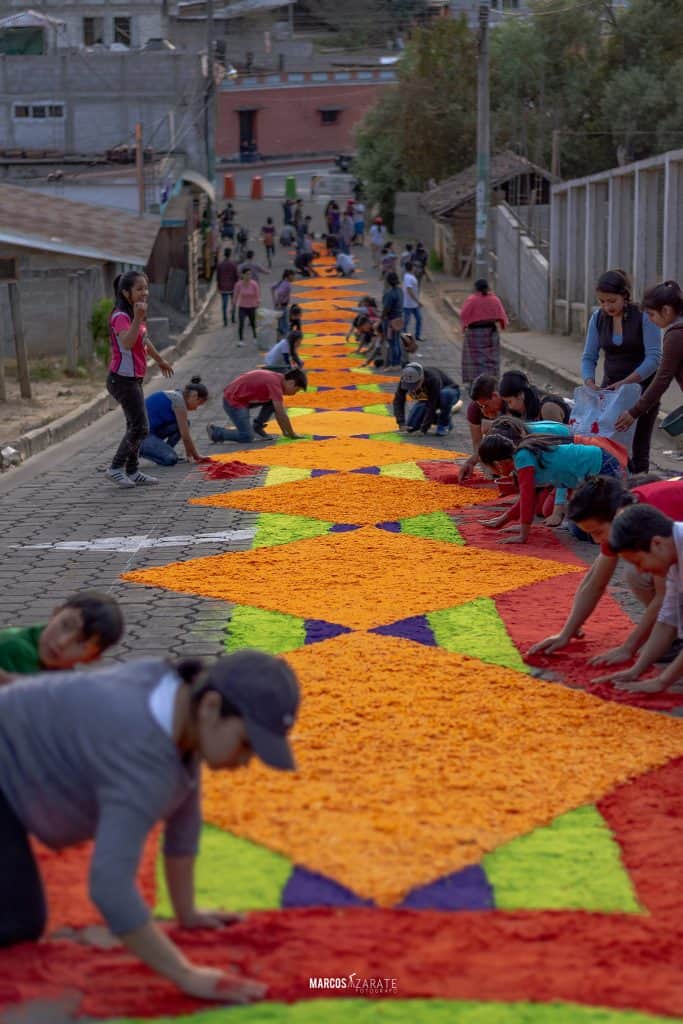 Momostenango amanece convertido en manos de alfombra y en alfombra de corazones, flores y plegarias – SoyMigrante.com REVISTA Colorido tapiz de flores elaborado por la comunidad en una calle de barrio en celebración cultural. Participación activa y tradición en evento artístico para fortalecer la identidad local. – SoyMigrante.com REVISTA