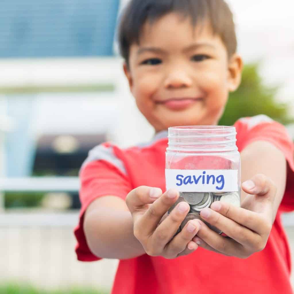 Ahorro, niño mostrando frasco con monedas y etiqueta "Saving" en inglés, promoviendo finanzas para niños y educación en ahorro en familia. – SoyMigrante.com REVISTA