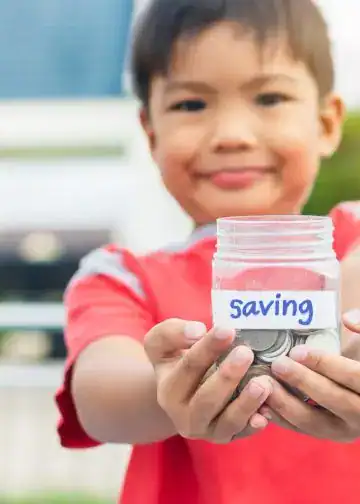 Alfabetización financiera para niños enseñar a ahorrar e invertir5 – SoyMigrante.com REVISTA Ahorro, niño mostrando frasco con monedas y etiqueta "Saving" en inglés, promoviendo finanzas para niños y educación en ahorro en familia. – SoyMigrante.com REVISTA