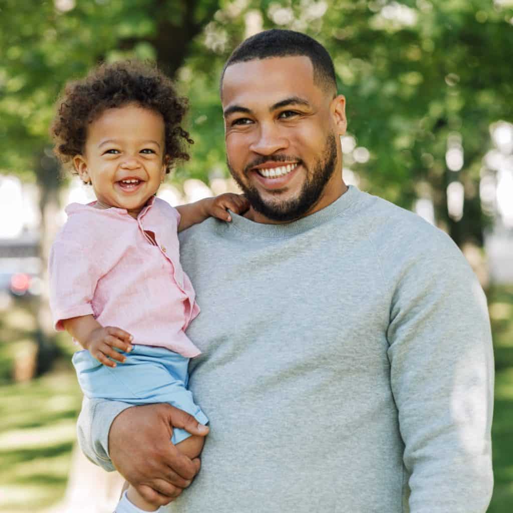 Padre e hija en parque, sonriendo, celebración de vínculos familiares y alegría. – SoyMigrante.com REVISTA