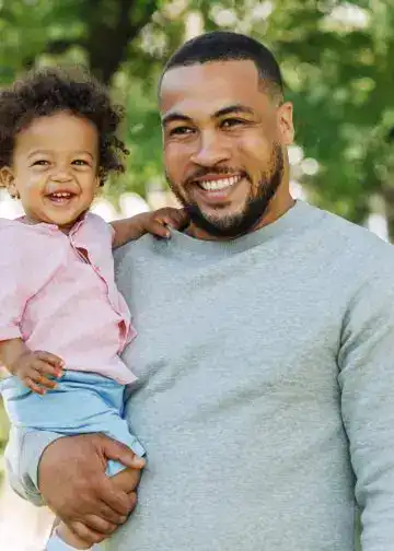 Padre e hija en parque, sonriendo, celebración de vínculos familiares y alegría. – SoyMigrante.com REVISTA