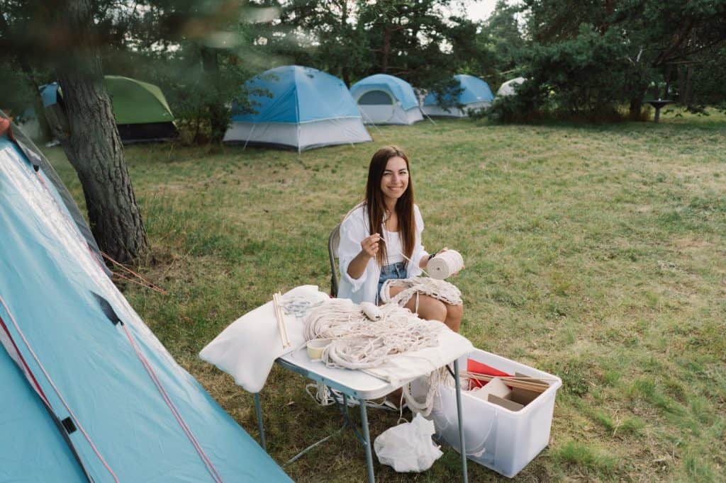 Mujer tejiendo en campamento con tiendas de campaña, escenario de historias migrantes y comunidad.