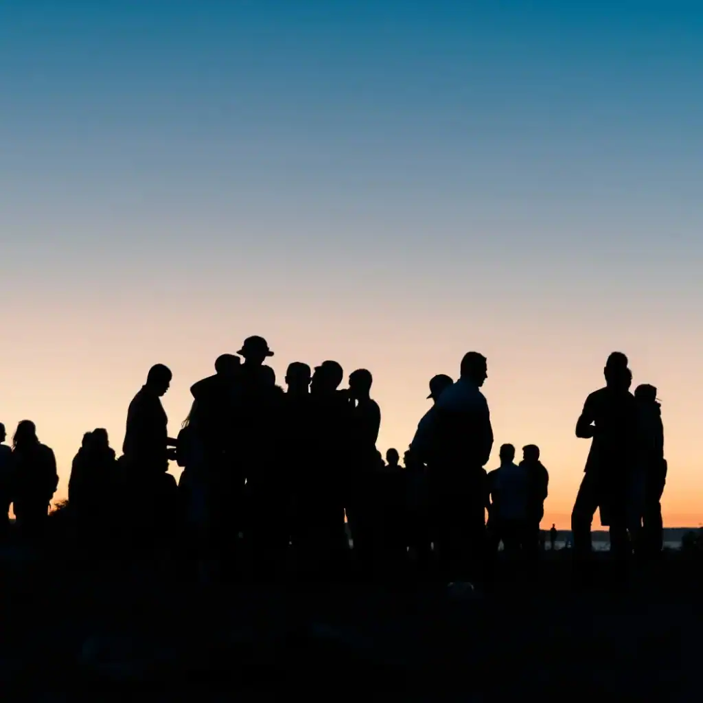 Personas migrantes en reunión al atardecer, siluetas en un entorno social, resaltando temas de migración, comunidad y cultura en la revista SoyMigrante.com REVISTA. – SoyMigrante.com REVISTA