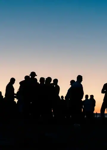 Personas migrantes en reunión al atardecer, siluetas en un entorno social, resaltando temas de migración, comunidad y cultura en la revista SoyMigrante.com REVISTA.