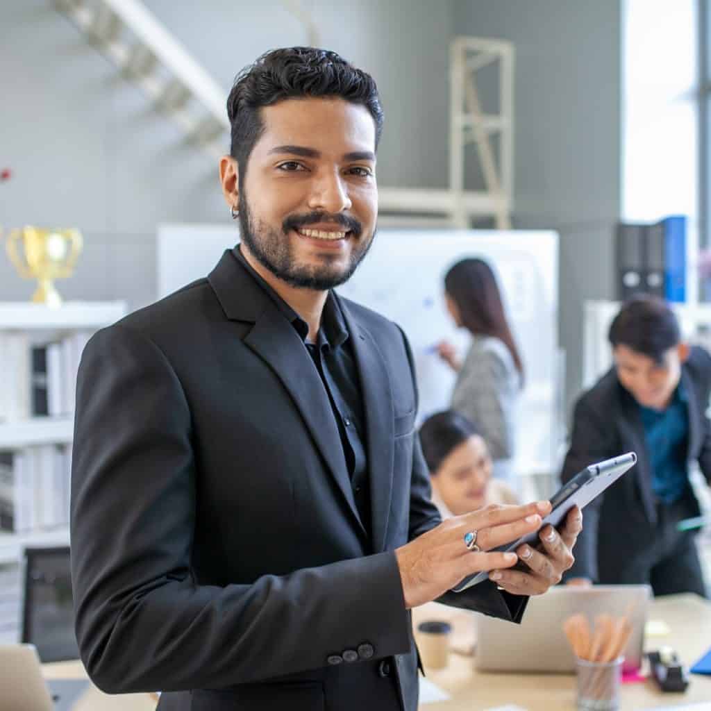 Joven profesional con barba en oficina moderna, usando traje y sosteniendo una tableta digital, rodeado de colegas trabajando en un ambiente corporativo y dinámico, promoviendo innovación y liderazgo. – SoyMigrante.com REVISTA