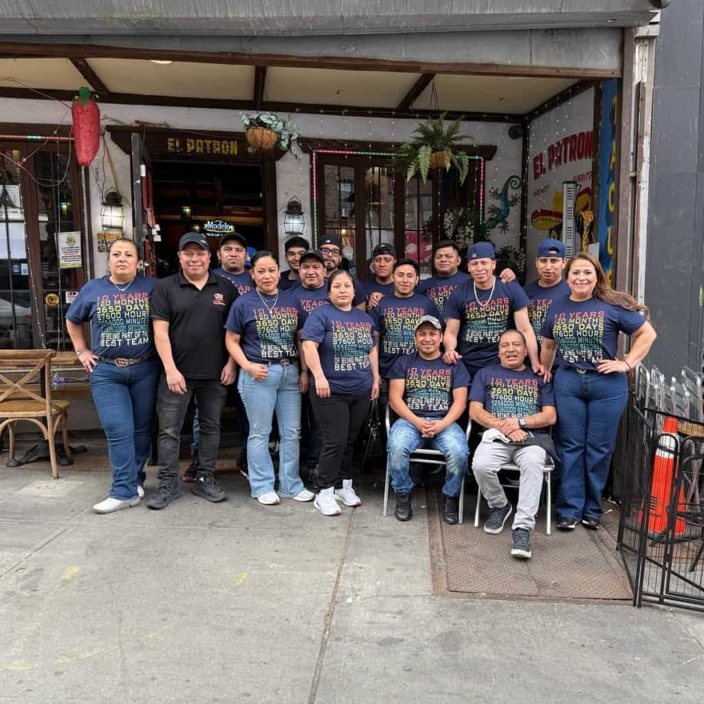 Grupo de migrantes y personal en la fachada de un restaurante en celebración del aniversario de SoyMigrante.com. Valorando la diversidad, comunidad y la historia de los inmigrantes en el país.