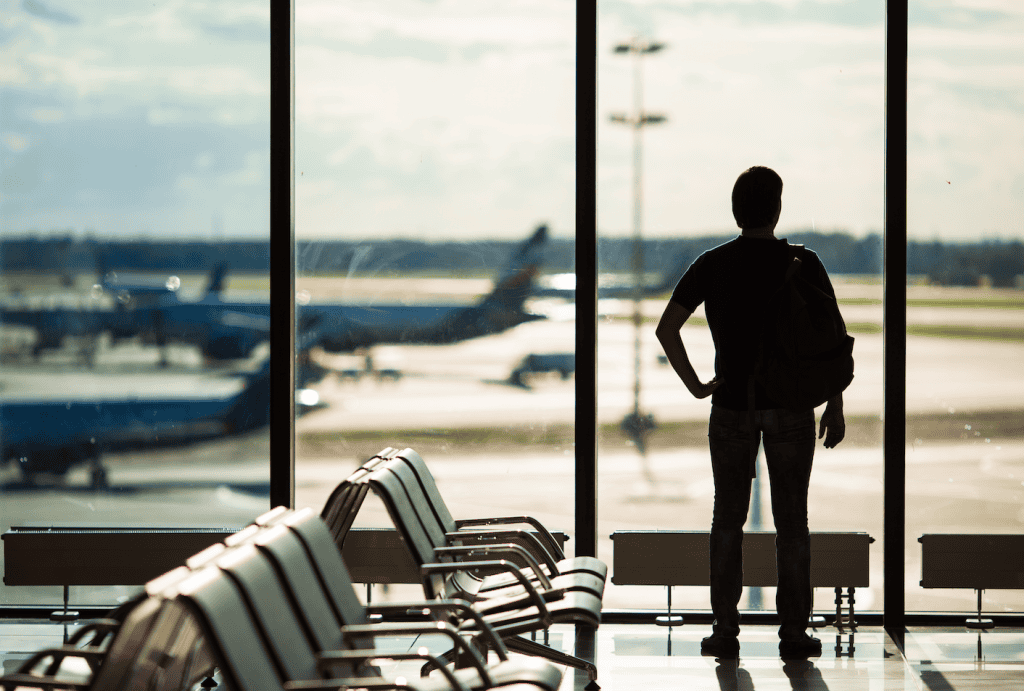 Archivo de espera en aeropuerto con vistas a aviones y pasajero con mochila, enfoque en viajes, migración y derechos de migrantes en contexto de revista SoyMigrante.com REVISTA.