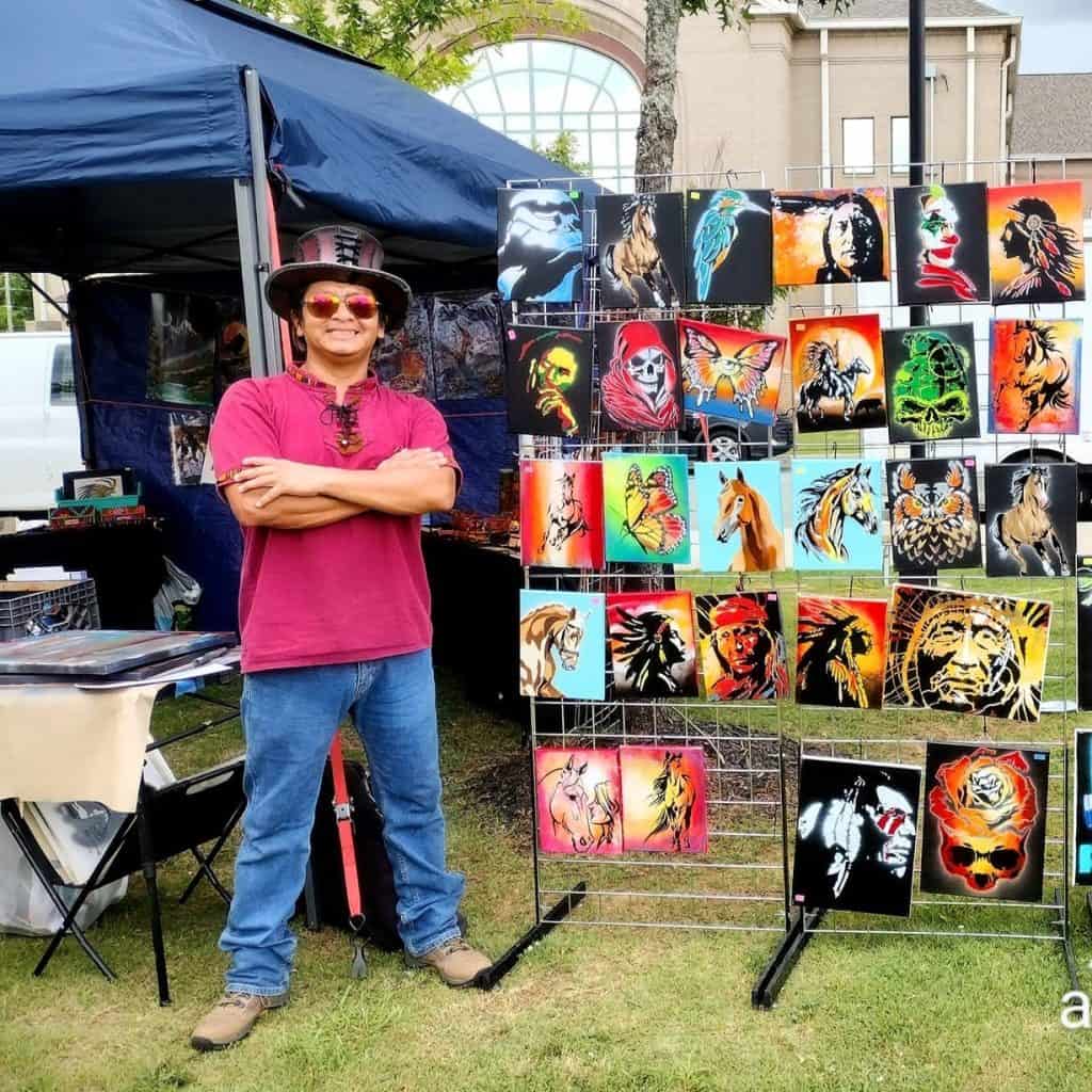 Colaborador de SoyMigrante exhibiendo arte de tatuajes y pinturas en mercado artesanal. Diseño y arte en lienzo, inspiración de migrantes y cultura en evento artístico. – SoyMigrante.com REVISTA