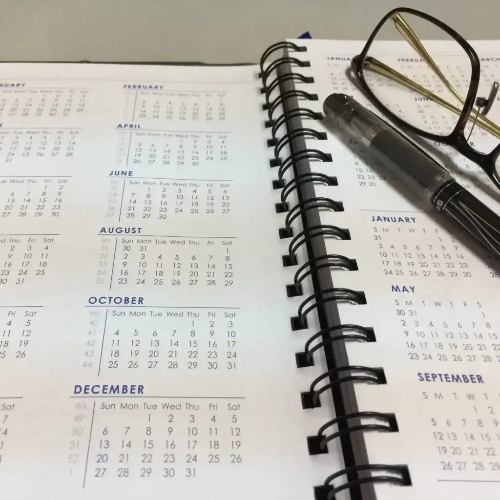 Calendario mensual en agenda con gafas, bolígrafo y lentes de lectura sobre papel, elementos de oficina, planificación, organización, productividad. – SoyMigrante.com REVISTA