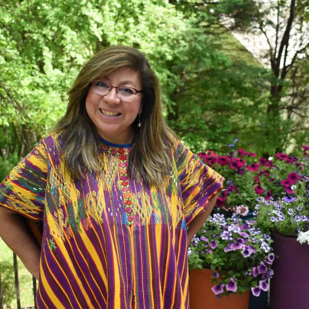 Mujer sonriente con vestimenta tradicional en un entorno natural, resaltando la cultura indígena y la diversidad en la comunidad migrante. – SoyMigrante.com REVISTA