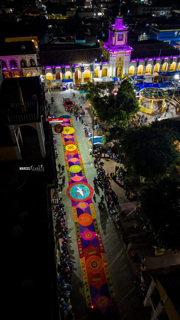 La noche cubre de sombras las calles, excepto aquellas donde pasa el Señor. – SoyMigrante.com REVISTA Coloridas calles iluminadas, feria y festividad en un centro histórico al atardecer, con gente disfrutando y decoración vibrante. – SoyMigrante.com REVISTA