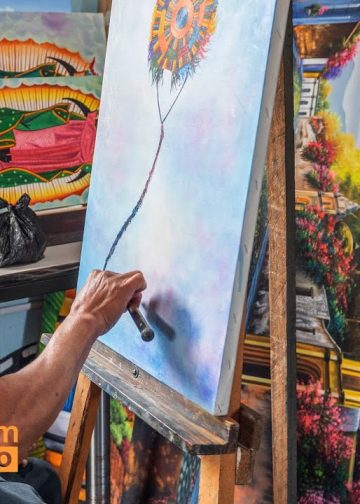 Migrant artist painting a blue-toned canvas at an easel, surrounded by colorful artworks; SoyMigrante.com MERCADO logo bottom left.