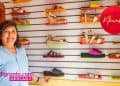 Smiling woman in a light blue shirt stands beside a wall display of colorful sandals in a shoe shop; SoyMigrante.com MERCADO logo visible.