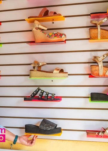 Smiling woman in a light blue shirt stands beside a wall display of colorful sandals in a shoe shop; SoyMigrante.com MERCADO logo visible.