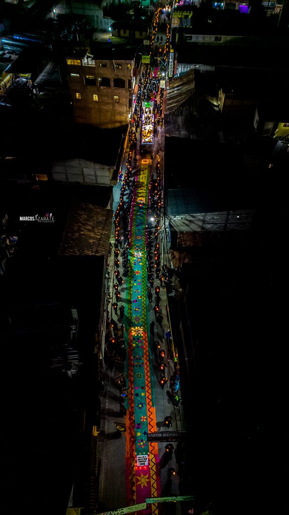 "Juntos como hermanos, miembros de la Iglesia, vamos caminando, al encuentro del Señor" – SoyMigrante.com REVISTA Paseo tradicional con coloridas alfombras de sal en la calle durante la celebración, iluminado por antorchas y luz artificial en un evento cultural nocturno en México. – SoyMigrante.com REVISTA