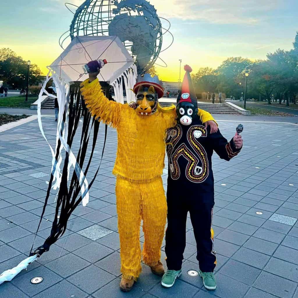 Hombre disfrazado de jaguar y otro con máscara de payaso en celebración tradicional mexicana en una plaza pública al atardecer. Personas y espacios abiertos en el fondo, ambiente festivo y cultural. – SoyMigrante.com REVISTA