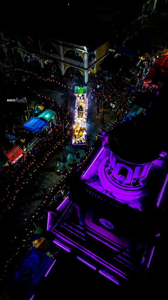 Una devoción vista desde el cielo: una ofrenda de corazones hermanos atraviesa el pueblo. El Santo Entierro regresa al templo parroquial de Momostenango. Ahora a esperar la Resurrección del Señor – SoyMigrante.com REVISTA Desfile festivo nocturno con iluminación vibrante en la calle principal, lleno de audiencia y alegría, celebración cultural, tradición, comunidad y música en un evento destacado. – SoyMigrante.com REVISTA