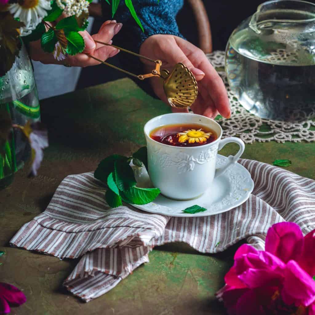 Copa de té con flor decorativa en taza blanca, rodeada de flores y plantas, en un ambiente acogedor y natural. – SoyMigrante.com REVISTA