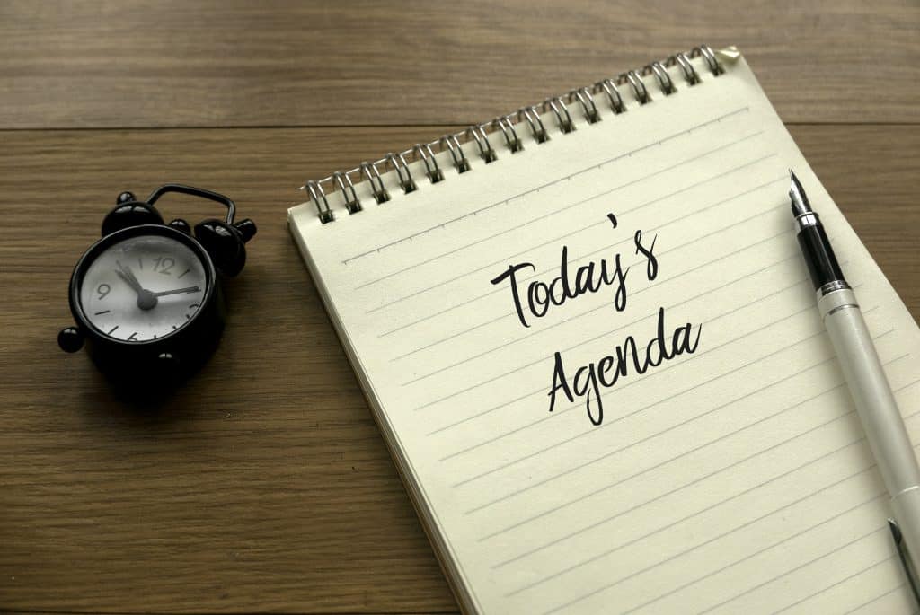 Reloj despertador y agenda con la frase "Today’s Agenda" en una mesa de madera, simbolizando planificación y organización para migrantes y comunidad hispana. – SoyMigrante.com REVISTA