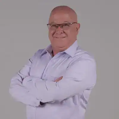 Hombre con gafas sonriendo, vestido con camisa blanca, en fondo gris, ilustración de confianza y profesionalismo para revista SoyMigrante.com REVISTA. – SoyMigrante.com REVISTA