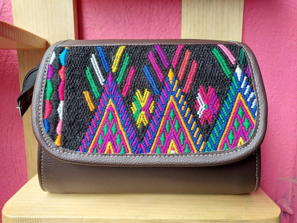 Colchón de tela tejida a mano con bordados coloridos en patrón tradicional indígena, sobre fondo negro y bordes en cuero marrón, exhibido en la revista SoyMigrante.com REVISTA.