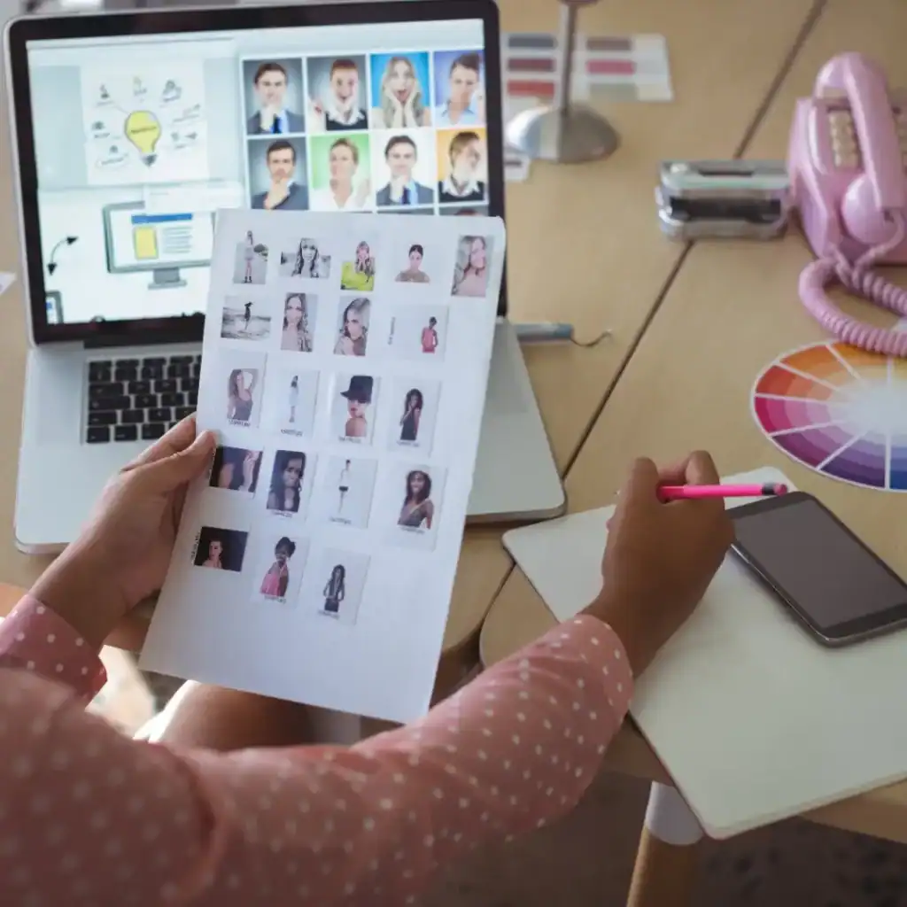 Retrato de un proceso de selección de fotos en una computadora portátil con una hoja de fotos en las manos, en un ambiente de oficina con elementos de diseño y comunicación. – SoyMigrante.com REVISTA