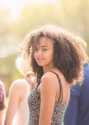 Chica joven con cabello rizado y sonrisa, en un entorno al aire libre con luz natural, representando temas de migración, juventud y diversidad en la comunidad latina.