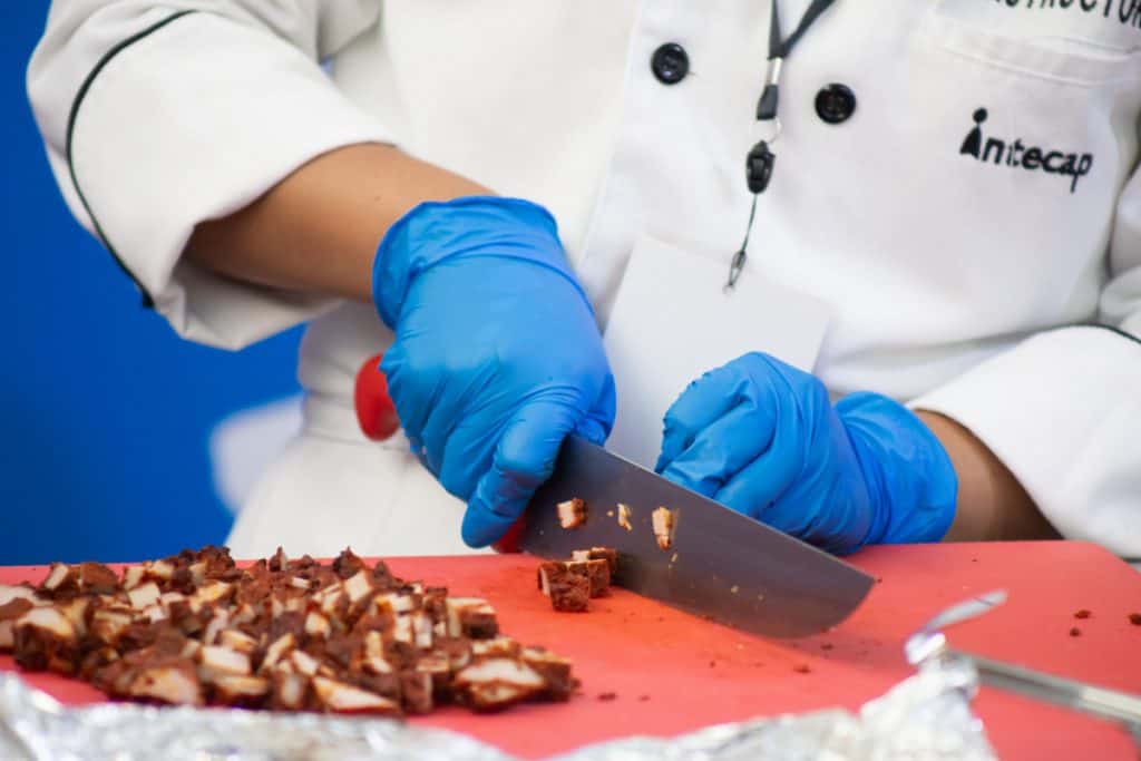 Cocinero con guantes azules cortando chocolate en una cocina profesional. – SoyMigrante.com REVISTA