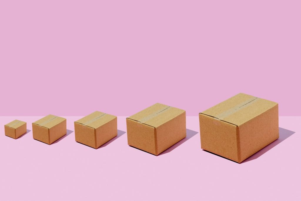 Alt: Cinco cajas de cartón tamaño diferente alineadas en una superficie rosa pastel, representando crecimiento o progresión. – SoyMigrante.com REVISTA