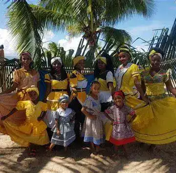Personas vestidas con trajes tradicionales en una celebración cultural en la playa, rodeadas de palmeras y un cielo despejado. Foto vibrante que destaca la identidad cultural y la festividad en un entorno paradisíaco. – SoyMigrante.com REVISTA